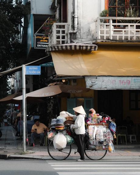 Ein Straßenhändler mit einem Fahrrad voller Waren wartet an einem Zebrastreifen vor einem gelben Gebäude.
