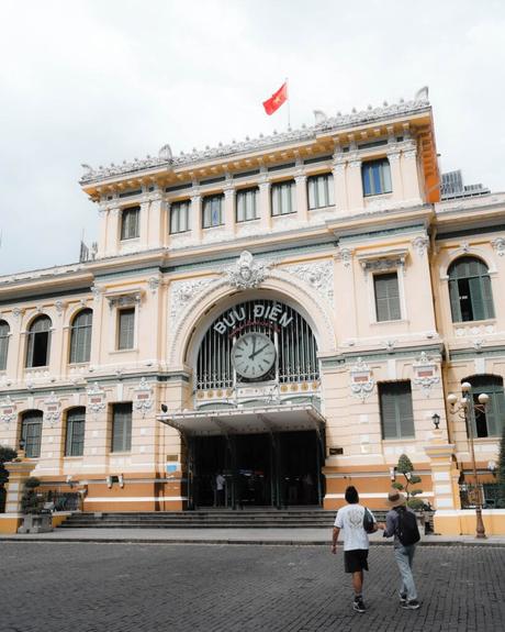 Zwei Personen gehen auf ein historisches gelbes Gebäude mit einer großen Uhr und der Aufschrift Buu Dien über dem Eingang zu.