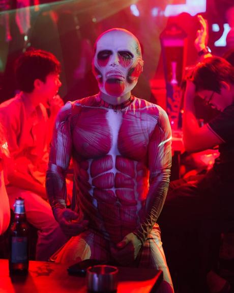 Person in einem Muskel- und Skelett-Bodysuit auf einer Party mit roter Beleuchtung, Menschen und Getränken im Hintergrund.