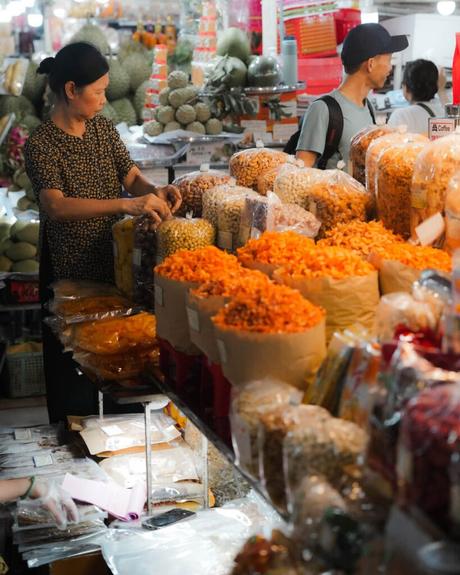 Eine Frau sortiert Trockenwaren an einem belebten Marktstand mit Tüten mit orangefarbenen und gelben Snacks.