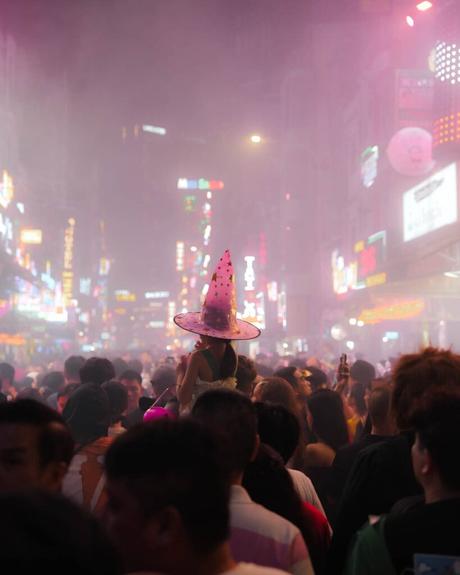 Eine Person mit einem rosafarbenen Zaubererhut fällt in einer belebten, neonbeleuchteten Stadtstraße bei Nacht und Nebel auf.