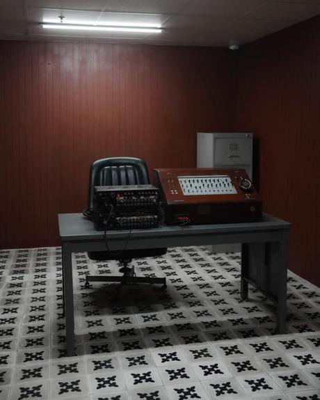 An einem Schreibtisch in einem spärlich eingerichteten Raum mit gemusterten Bodenfliesen stehen ein altes Bedienfeld und ein Stuhl.