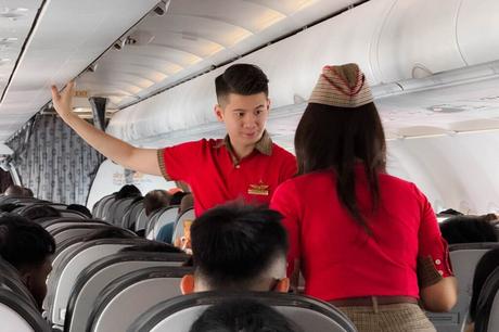 Zwei Flugbegleiterinnen in roten Uniformen unterhalten sich in einem Gang eines Flugzeugs zwischen sitzenden Passagieren.