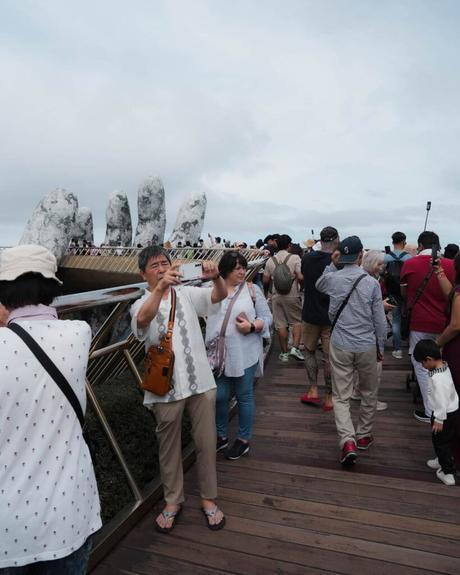 Touristen machen Fotos auf einer überfüllten Holzbrücke mit großen steinernen Handskulpturen im Hintergrund.