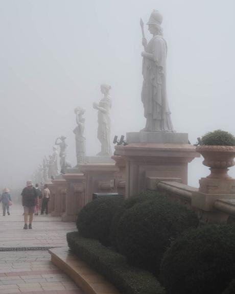 Die Menschen spazieren über eine neblige Terrasse, die von großen klassischen Statuen und gepflegten Büschen gesäumt ist.