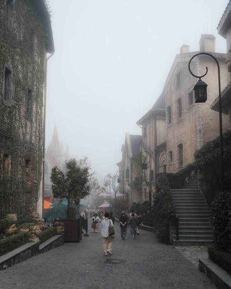 Die Menschen gehen eine neblige, kopfsteingepflasterte Straße entlang, die von alten Steinhäusern und Grünanlagen gesäumt ist.