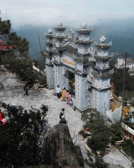 Verziertes Steintor an einem Tempel auf einem Berggipfel mit spazierenden Menschen und nebligen Bergen im Hintergrund.