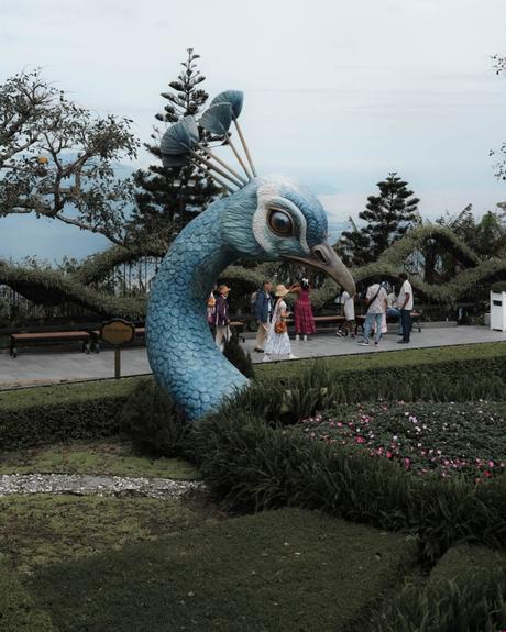 Eine große blaue Pfauenskulptur in einem Garten, in dem Menschen stehen und gehen.
