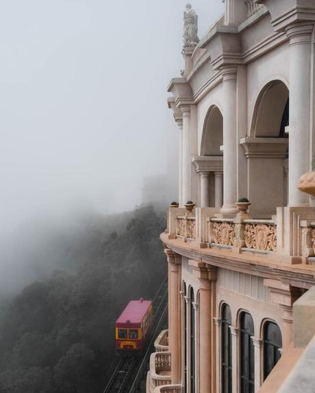 Ein rot-gelber Zug fährt durch den Nebel an einem verschnörkelten Gebäude mit Bögen und Statuen vorbei.