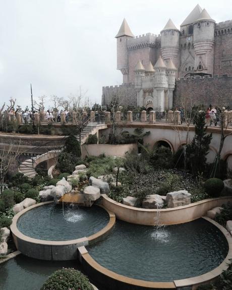 Ein märchenhaftes Schloss mit Gärten, Brunnen und Steinwegen unter einem wolkenverhangenen Himmel.