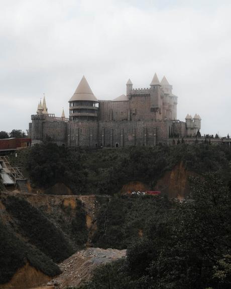 Mittelalterliche Steinburg mit Türmen auf einem Hügel, umgeben von Bäumen unter einem bewölkten Himmel.