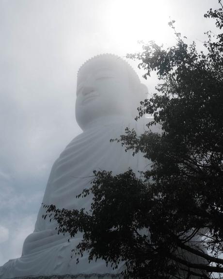 Große weiße Buddha-Statue hinter silhouettierten Baumzweigen, beleuchtet vom Sonnenlicht vor einem bewölkten Himmel.