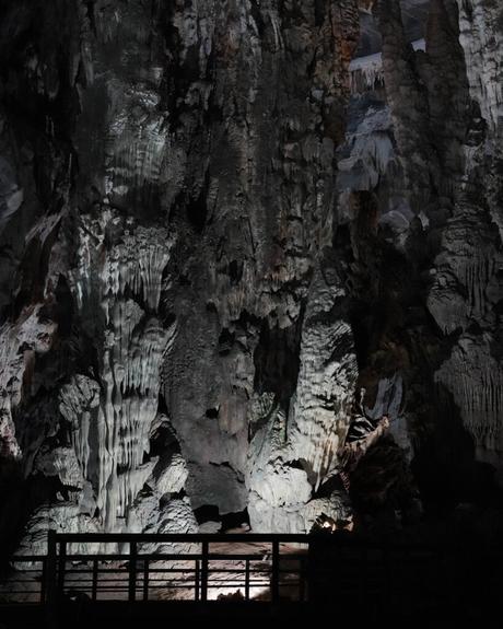 Schummerig beleuchtete Höhlenwand mit dramatischen Felsformationen und einem Holzgeländer im Vordergrund.