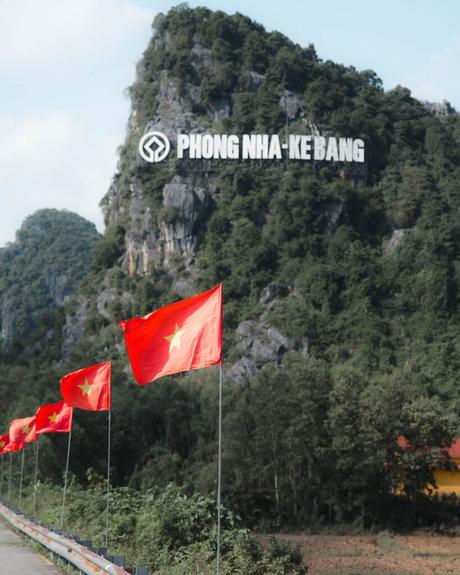 Vietnamesische Flaggen säumen eine Straße mit dem Schild Phong Nha - Ke Bang auf einem üppig grünen Berg im Hintergrund.