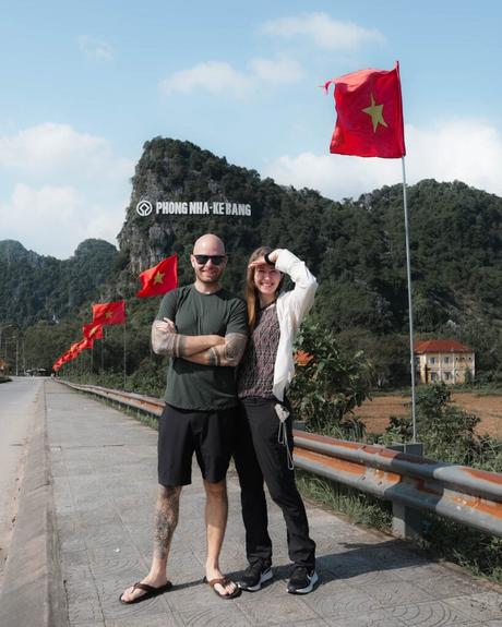 Zwei Personen posieren am Straßenrand mit vietnamesischen Flaggen und Bergen mit der Aufschrift Phong Nha-Ke Bang im Hintergrund.