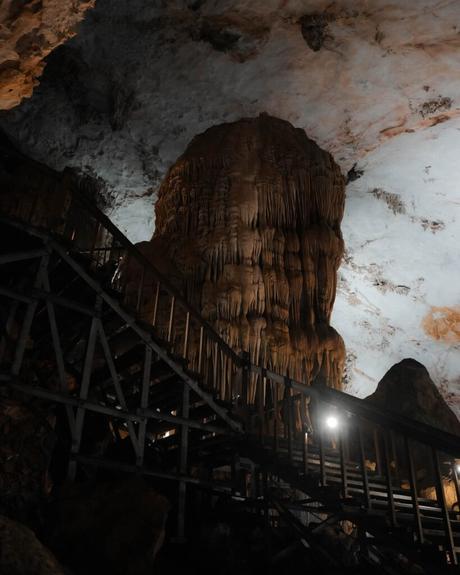 Ein großer Stalagmit erhebt sich neben einer Holztreppe in einer schwach beleuchteten Höhle mit einer strukturierten Decke.