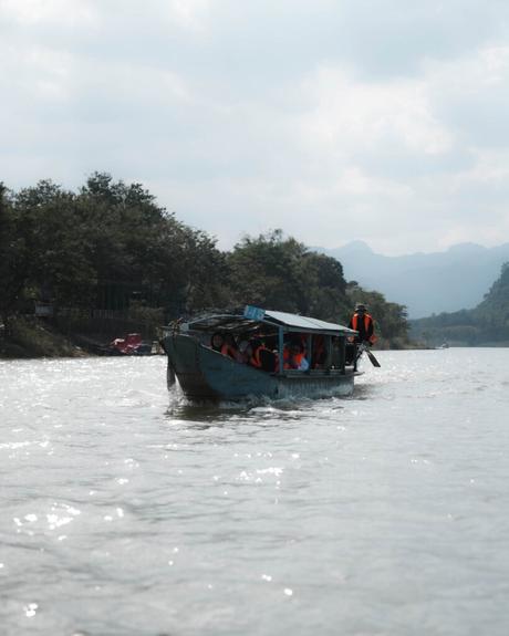 Ein blaues Boot mit Passagieren in Schwimmwesten fährt auf einem breiten Fluss, der von Bäumen und Hügeln umgeben ist.