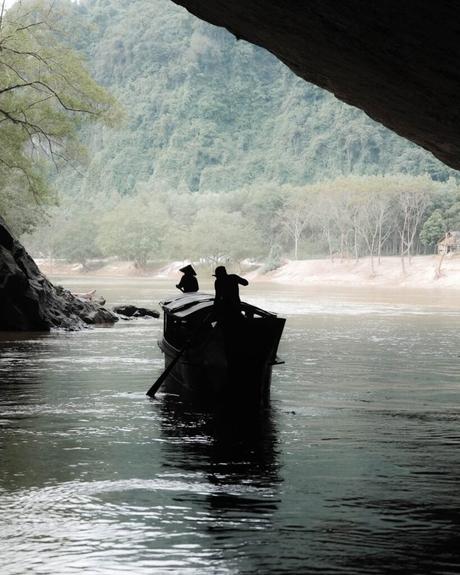 Silhouette von zwei Personen, die ein Boot auf einem Fluss rudern und aus einer Höhle in eine bewaldete Landschaft fahren.
