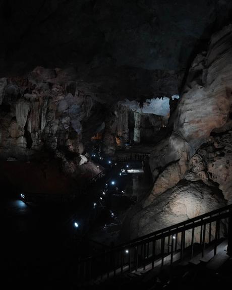 Das Innere der Höhle ist schwach beleuchtet, mit felsigen Wänden, einem Steg und kleinen Lichtern, die den Weg durch die Dunkelheit weisen.