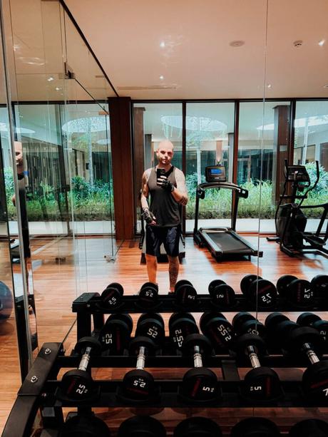 Ein Mann macht ein Selfie in einem Fitnessstudio mit Hanteln, Laufband und Trainingsgeräten, die sich in Spiegeln spiegeln.