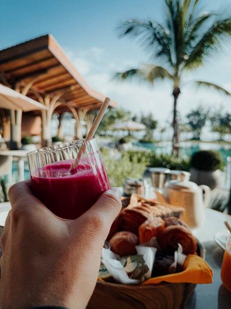 Eine Hand hält ein Glas roten Saft bei einem Frühstück am Pool mit Gebäck, Palme und Resort im Hintergrund.