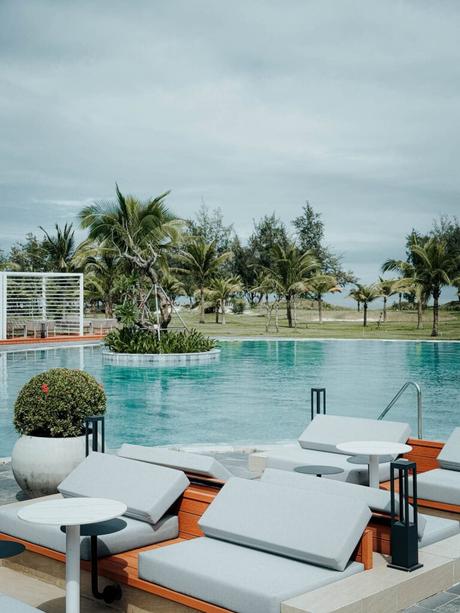 Liegestühle an einem Pool mit Palmen und Grünpflanzen unter einem bewölkten Himmel.