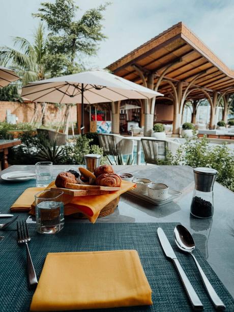 Frühstückstisch im Freien mit Gebäck, Getränken und Geschirr in einer Gartenterrasse mit Sonnenschirmen und Grünpflanzen.