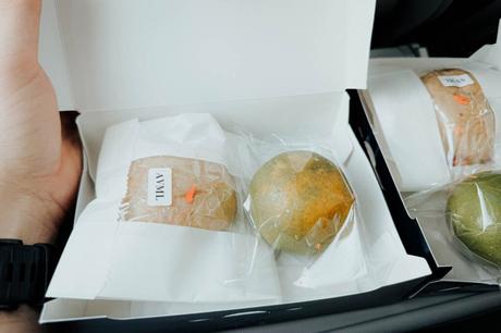 Eine Hand hält eine offene Schachtel mit zwei eingewickelten Backwaren, eine davon mit der Aufschrift TUAN, neben einer anderen geschlossenen Schachtel.