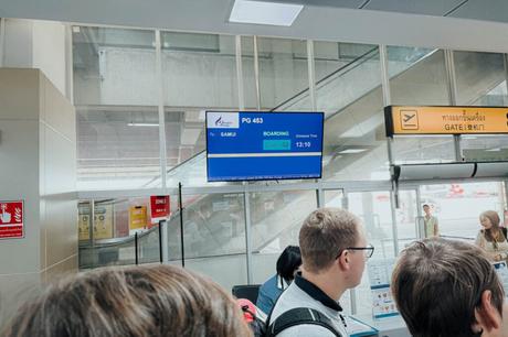 Menschen warten an einem Flughafen-Gate mit einem Bildschirm, auf dem die Boarding-Informationen für einen Flug nach Samui angezeigt werden.