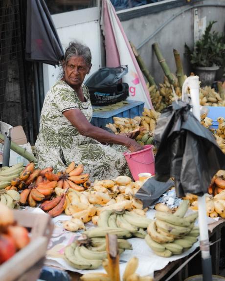 Ältere Frau in einem grünen Kleid verkauft verschiedene Bananen an einem Marktstand im Freien.