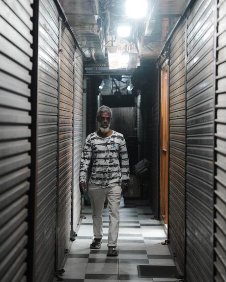 Ein Mann in einem gemusterten Hemd geht einen schmalen, schwach beleuchteten Korridor mit geschlossenen Metallläden entlang.