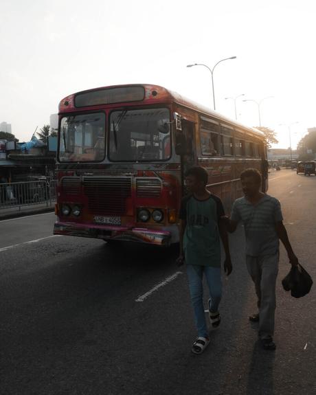 Zwei Männer gehen bei Sonnenuntergang auf einer Stadtstraße in der Nähe eines roten Busses spazieren, mit Verkehr und Straßenbeleuchtung im Hintergrund.