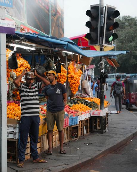 Zwei Männer stehen an einem Obststand mit Orangen und Melonen an einer belebten Straße unter einer Ampel.