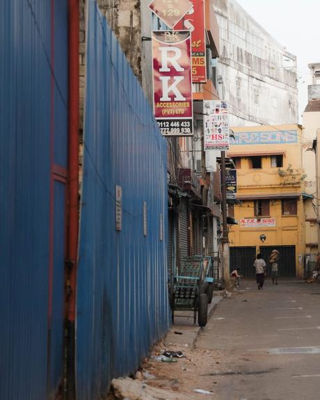 Schmale Gasse mit blauem Zaun, Ladenschildern und zwei Personen, die in der Ferne in der Nähe gelber Gebäude spazieren gehen.