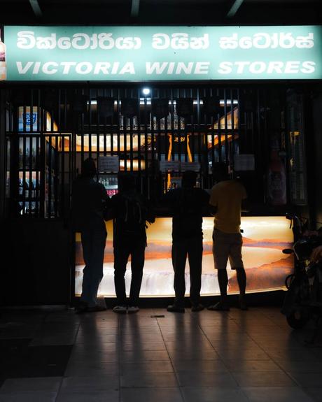 Vier Personen stehen nachts an der Theke des Victoria Wine Stores, zu sehen als Silhouette unter einem hellen Schild.
