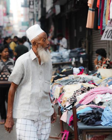 Ein älterer Mann geht auf einem belebten Markt an den Ständen vorbei, an denen Kleidung verkauft wird.