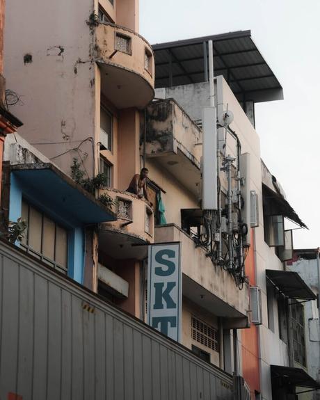 Ein Mann steht auf einem kleinen Balkon eines alten, verwitterten Wohnhauses, an dessen Außenwand Antennen angebracht sind.