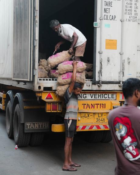 Ein Mann entlädt Säcke von einem Lastwagen, während eine andere Person einen Sack darunter hält; eine dritte Person ist in der Nähe.