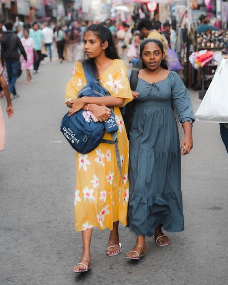 Zwei junge Frauen in langen Kleidern gehen zusammen auf einer belebten Straße, die von Geschäften und Menschen gesäumt ist.