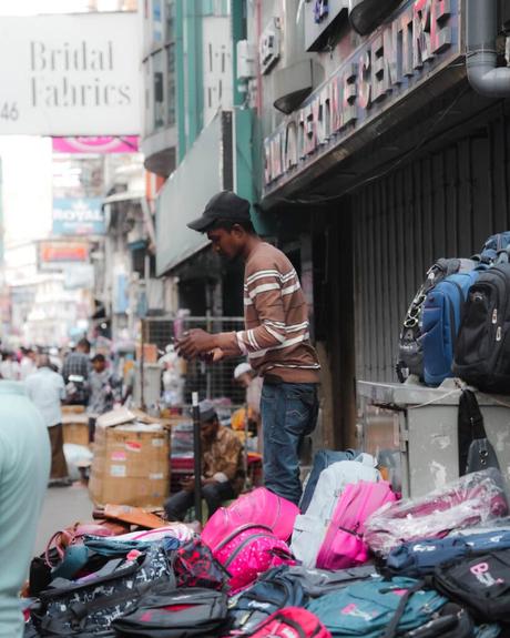Ein Straßenverkäufer steht neben einer Auslage mit bunten Rucksäcken auf einem belebten städtischen Bürgersteig.