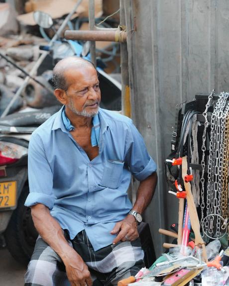 Ein älterer Mann in einem blauen Hemd sitzt an einem Straßenstand, an dem verschiedene Metallwerkzeuge und Ketten verkauft werden.