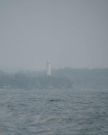 Ein weißer Leuchtturm steht an einer nebligen Küste, mit Wellen im Vordergrund und Bäumen im Hintergrund.