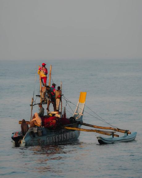 Ein kleines Fischerboot mit fünf Personen auf ruhigem Wasser, eine Person steht hoch und hält einen großen Fisch.