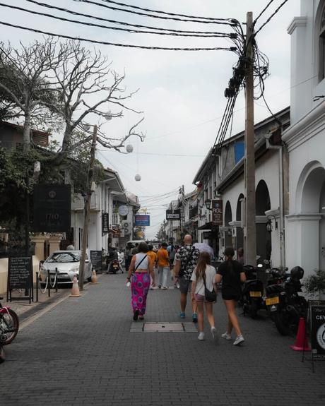 Unter einem wolkenverhangenen Himmel gehen Menschen eine Kopfsteinpflasterstraße entlang, die von Geschäften und Motorrollern gesäumt ist.