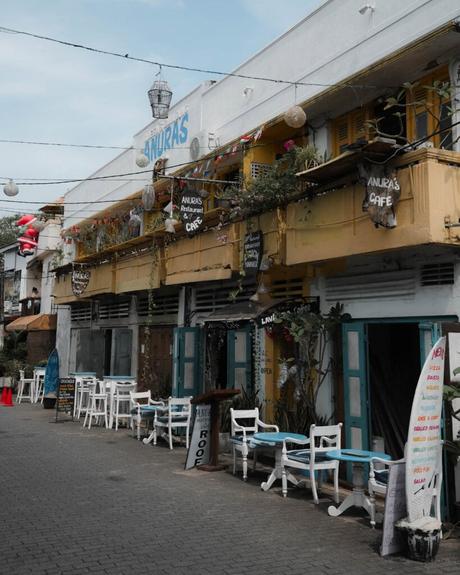 Außenansicht eines bunten Cafés mit blauen Stühlen, Tischen, Pflanzen und handgeschriebenen Schildern in einer belebten Straße.
