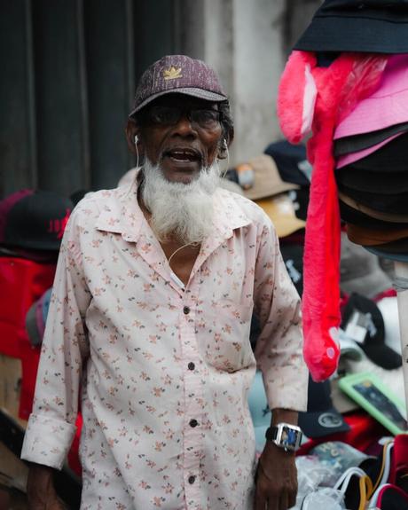 Älterer Mann mit langem weißem Bart und Brille, der mit Hüten auf einem Markt im Freien steht und freundlich lächelt.