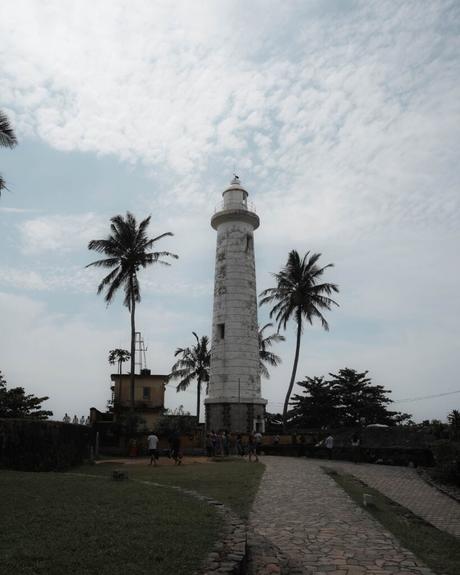 Ein hoher weißer Leuchtturm steht inmitten von Palmen und Menschen, die unter einem wolkenverhangenen Himmel auf einem Steinweg spazieren gehen.