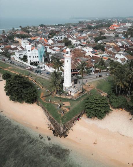 Luftaufnahme eines weißen Leuchtturms in der Nähe eines Sandstrands, umgeben von Grünflächen und einer Küstenstadt.