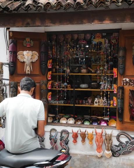 Ein Mann auf einem Motorroller betrachtet ein lebendiges Schaufenster mit Masken und buntem Kunsthandwerk.