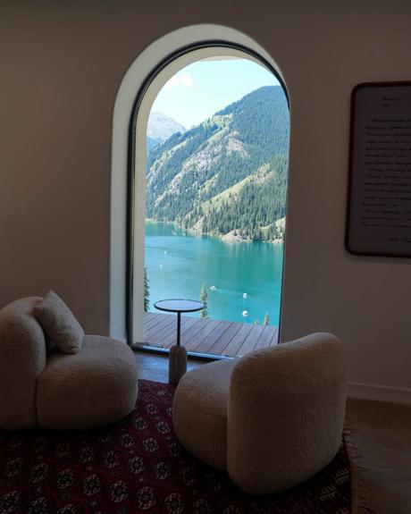 Zwei gemütliche Stühle und ein Tisch stehen vor einem Fenster mit Blick auf einen Bergsee, hinter dem sich Bäume und Hügel erstrecken.